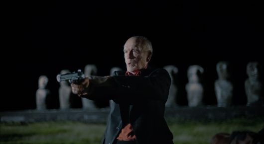 Still from Wild Horse Nine: an older man in dark clothes aims a pistol in a dark room, with a row of white busts on shelves behind him.