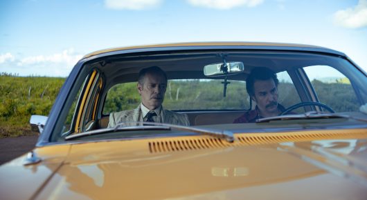 Still from Wild Horse Nine: two men sit in a vintage yellow and blue car, seen through the windscreen on a country road.