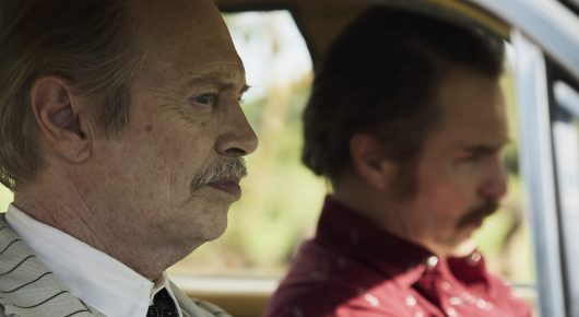 Still from Wild Horse Nine: a close view of two men in the front of a car, an older man with a grey moustache in a striped shirt and a younger man with a dark moustache in a red shirt.