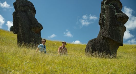 Still from Wild Horse Nine: two people stand in a grassy field, dwarfed by two large carved stone heads.