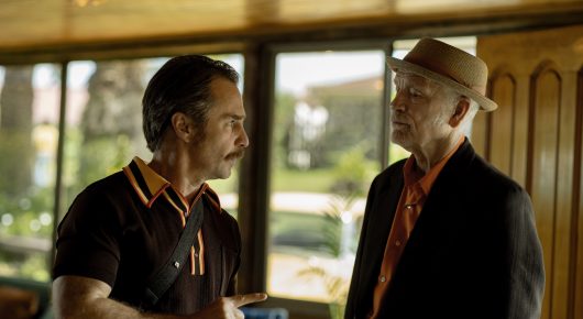 Still from Wild Horse Nine: two men talk in a wood-panelled room, one in a dark polo shirt and the other in a hat and dark jacket.