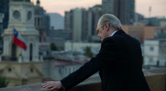 Still from Wild Horse Nine: an older man in a dark suit leans on a balcony wall overlooking a city, with a red, white and blue flag on a rooftop.