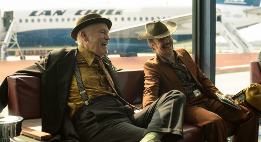 Still from Wild Horse Nine: two men in hats and long coats sit on a sofa in an airport lounge, with an aeroplane visible through the window.