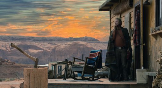Still from Wild Horse Nine: a figure stands on a wooden porch looking out over hills at sunset, with a chopping block and an axe in the foreground.