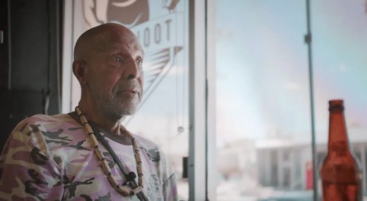 Still from Welcome to Tool Shed: an older man with a shaved head and a beaded necklace, wearing a pink floral shirt, looks to one side beside a window and a beer bottle.