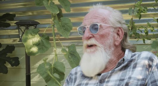 Still from Welcome to Tool Shed: an older man with a long white beard and tinted glasses looks to one side in front of green plants.