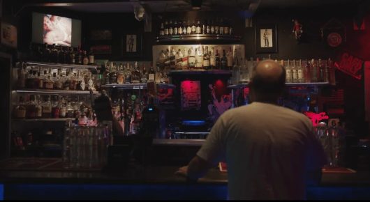 Still from Welcome to Tool Shed: a man seen from behind stands at a dimly lit bar lined with bottles and memorabilia.