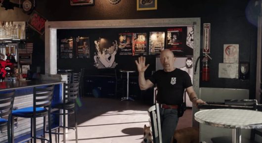 Still from Welcome to Tool Shed: a man in a black T-shirt waves from the middle of a bar and music venue hung with posters.