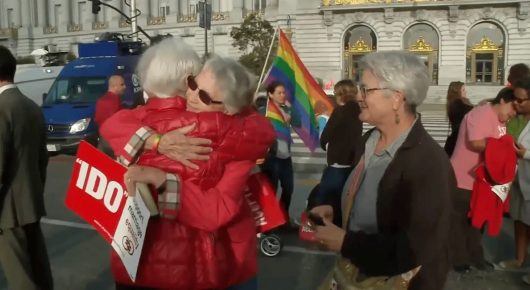 Still from Welcome to Tool Shed: older people hug at a street protest, one holding a red sign, with a rainbow flag and a large building behind them.