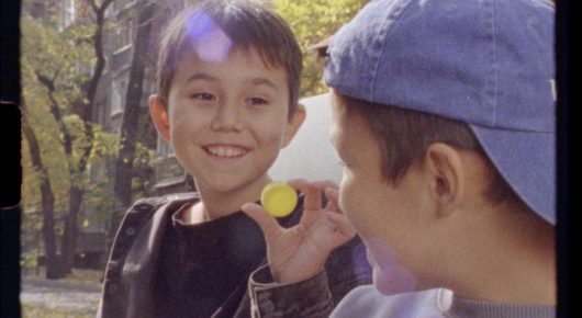 Still from Aryk: a smiling boy looks at the camera while a boy in a blue cap is in the foreground, on a tree-lined street.