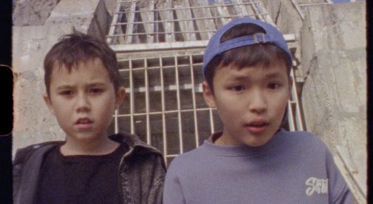 Still from Aryk: two boys stand side by side outdoors in front of a metal gate, one in a blue cap and one in a dark jacket.