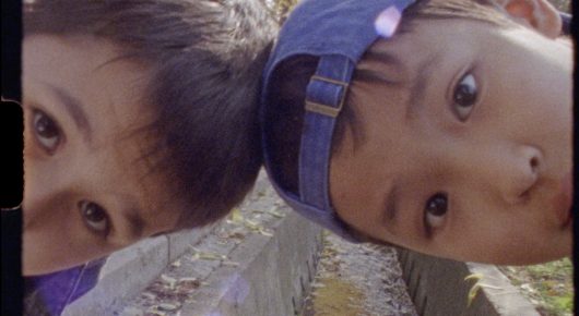 Still from Aryk: a close view of two boys looking down at the camera, one in a blue cap, with gravel behind them.