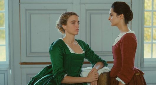 Still from Portrait of a Lady on Fire: two women in period dress, one seated in green and one standing in red, face each other in a bright room with white panelled doors.