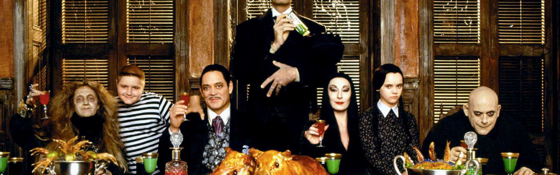 Still from Addams Family Values: the Addams family sit around a long table laden with food in a dark wood-panelled dining room.