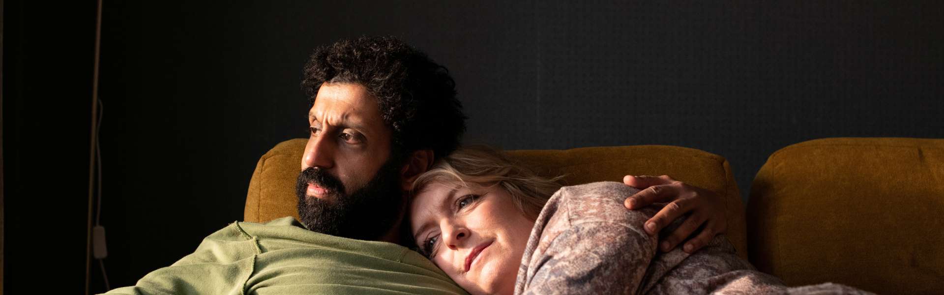Still from Ali & Ava: a bearded man in a green jumper and a woman lie close together on a yellow sofa in a dim room.