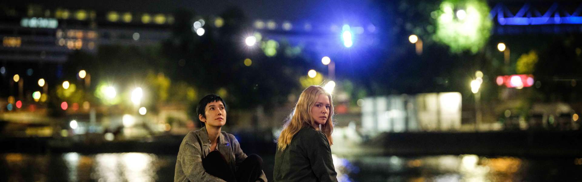 Still from All of a Sudden: two people sit on a stone quayside at night, with city lights reflected in the river behind them.