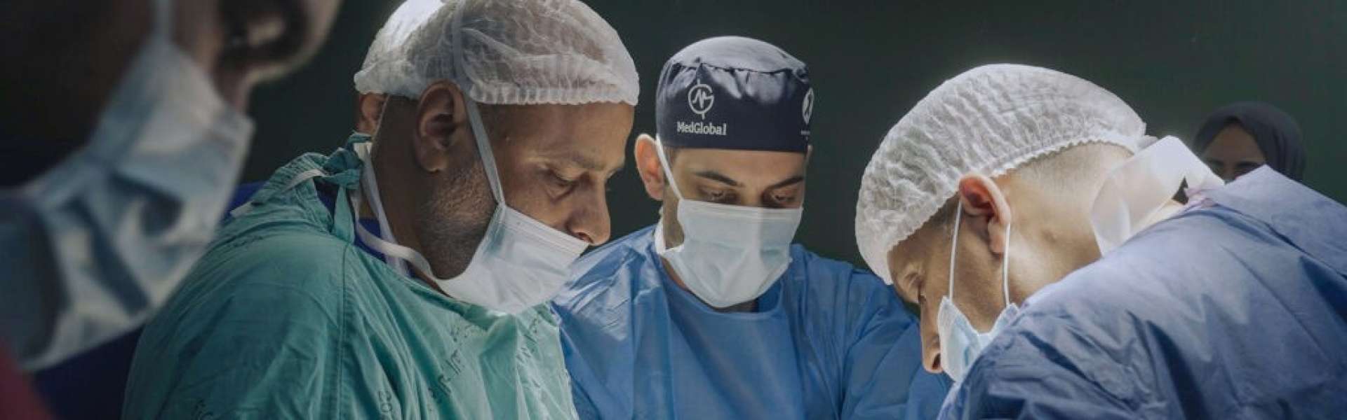 Still from American Doctor: surgeons in scrubs, masks and hair nets work over an operating table under bright lights.