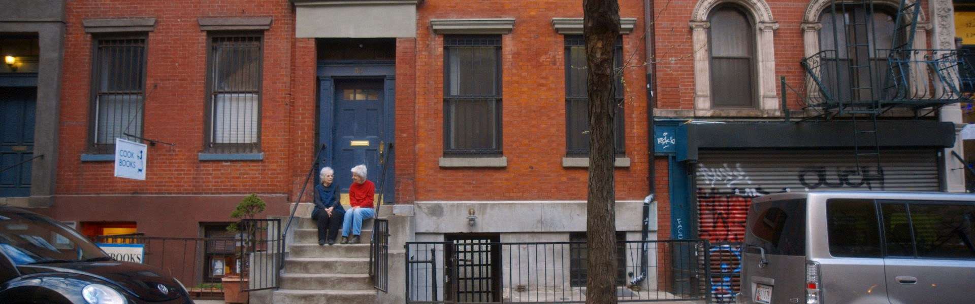 Still from Artists in Residence: a red-brick townhouse street with parked cars and a van, with steps up to a blue front door.