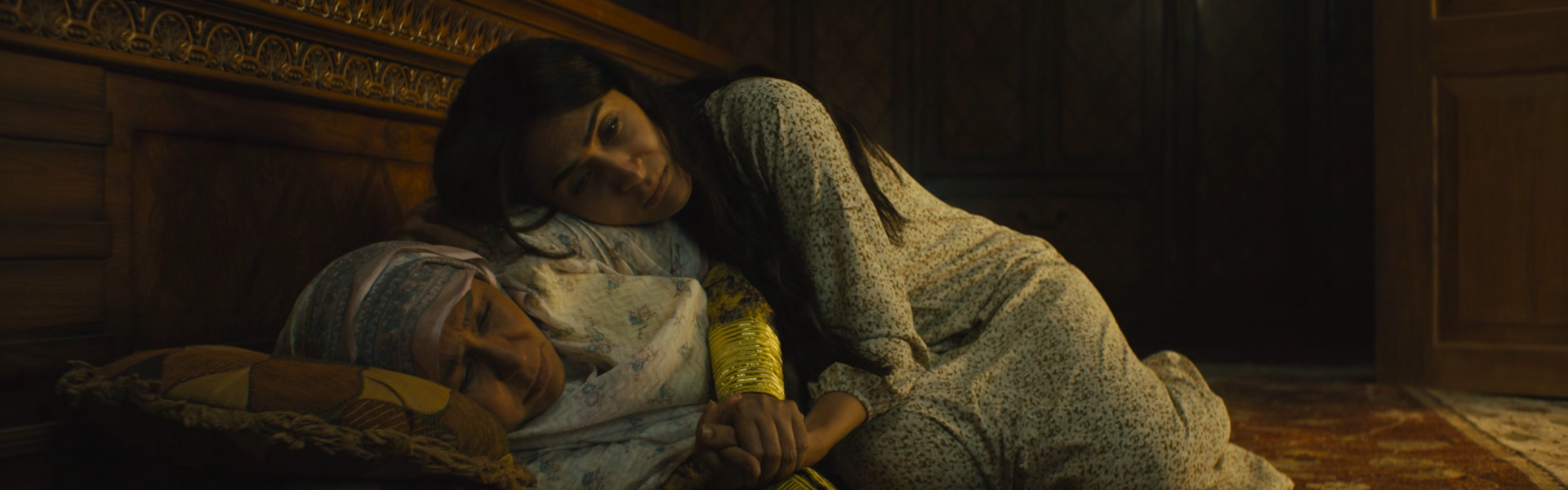 Still from Baab: two people in pale clothes lie close together on a rug and cushions in a dim, wood-panelled room.