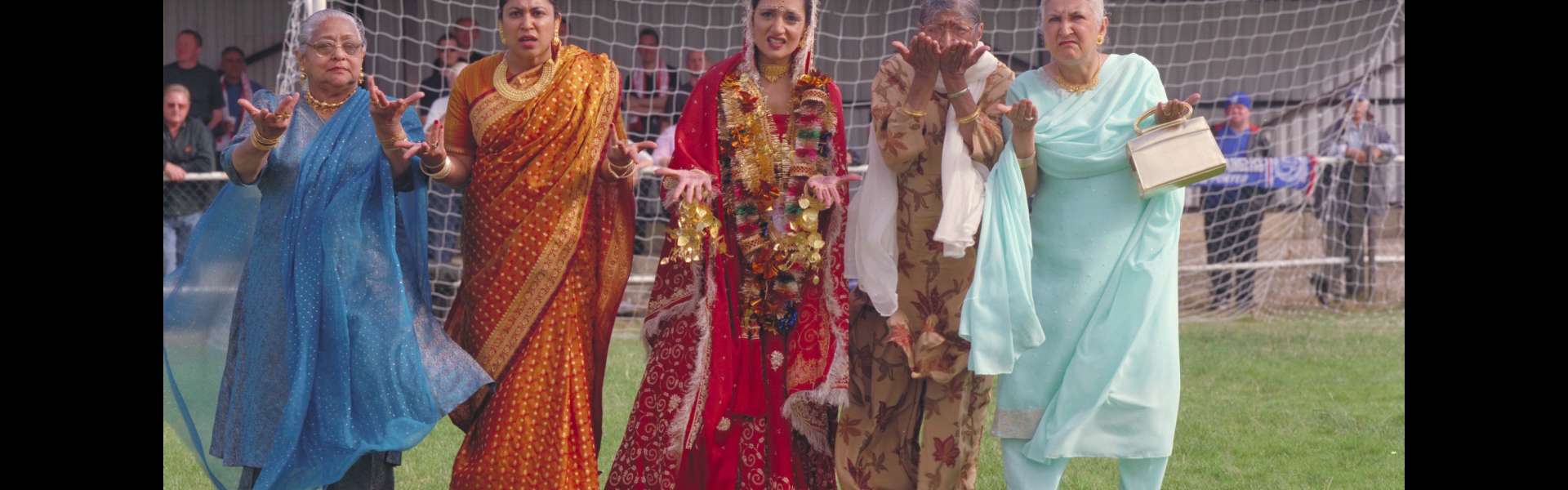 Still from Bend It Like Beckham: five women in brightly coloured saris and salwar kameez stand in front of a football goal on a pitch.