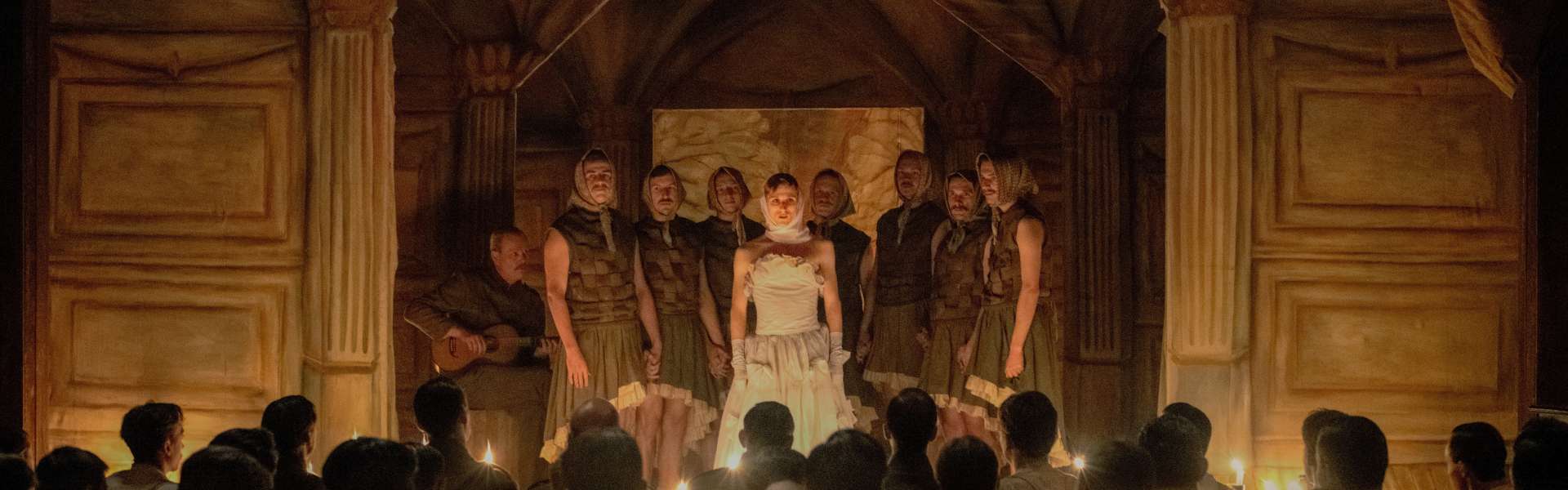 Still from Coward: a crowd of soldiers watches a candlelit stage where a few performers stand in front of ornate columns.