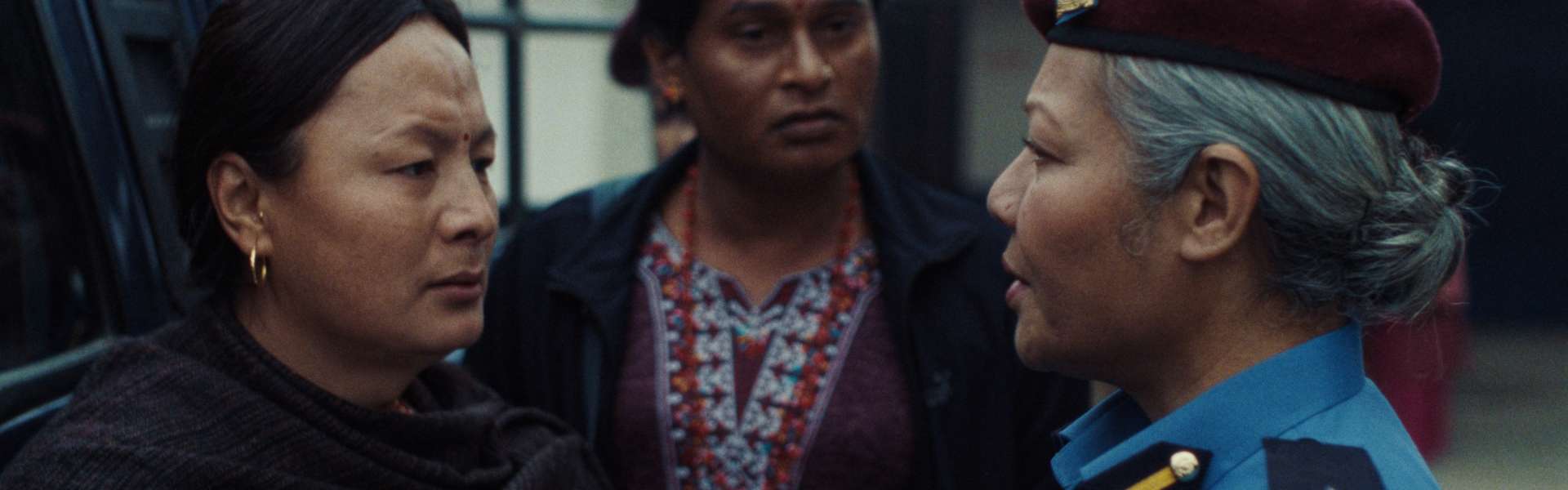 Still from Elephants in the Fog: three women stand close together on a platform, one in a maroon beret and blue uniform, one in a beaded necklace and one in a black shawl.