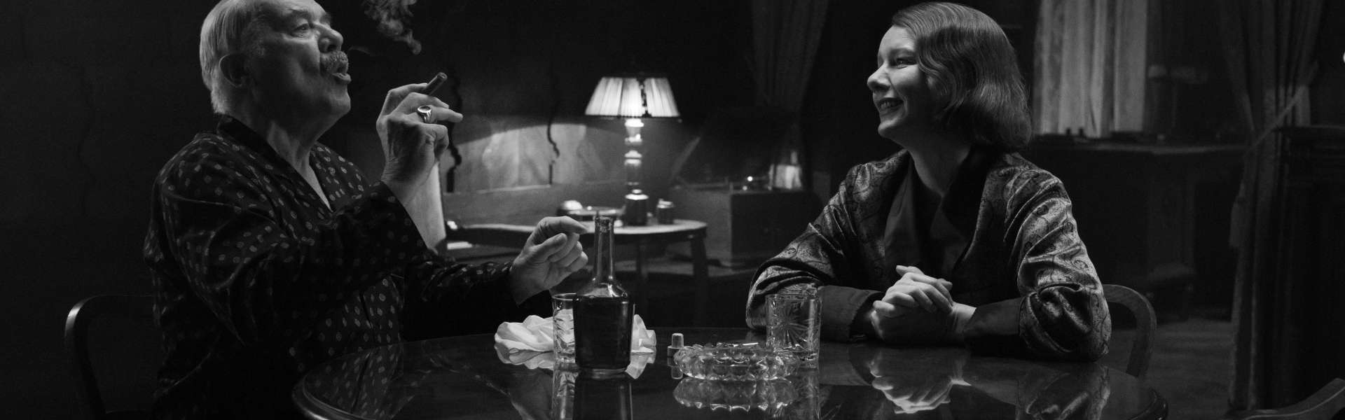 Still from Fatherland: a man smokes a cigarette at a round table opposite a woman in a dim café with a lamp, in black and white.