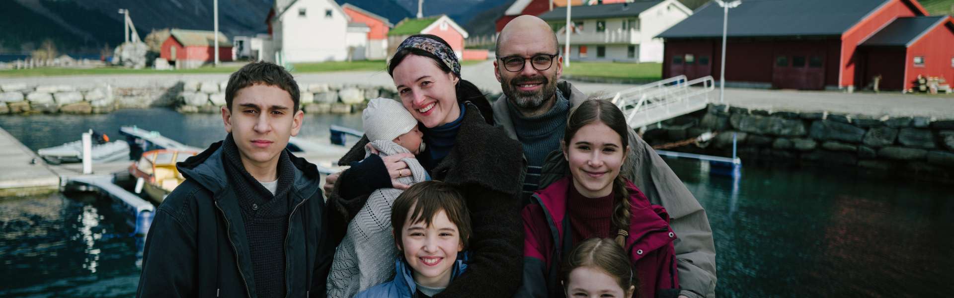Still from Fjord: a family of six pose together in front of a fjord village with red and white buildings and mountains.