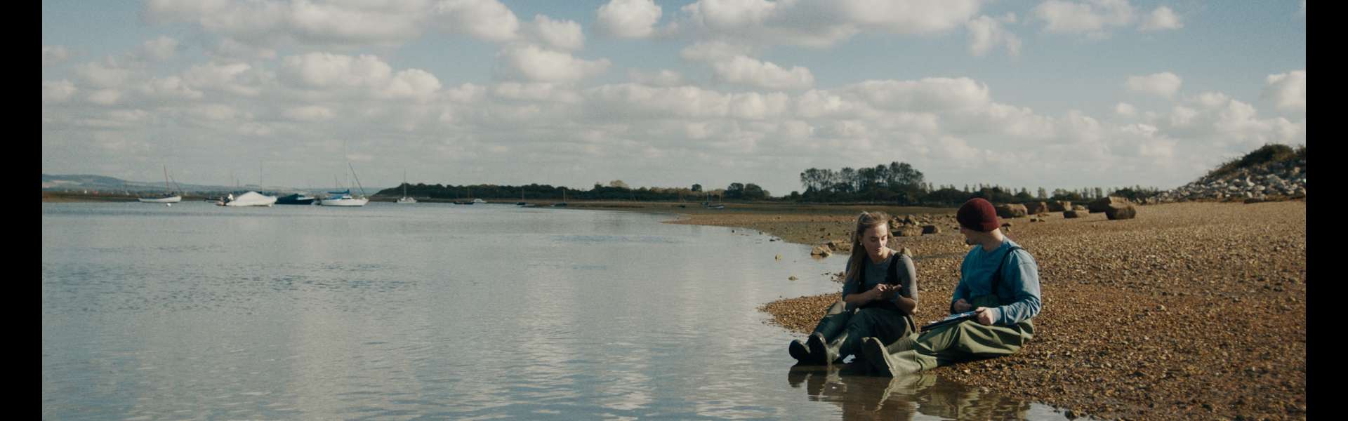 Still from Green Grass: two people sit on the shore of a calm estuary under a blue sky with clouds.
