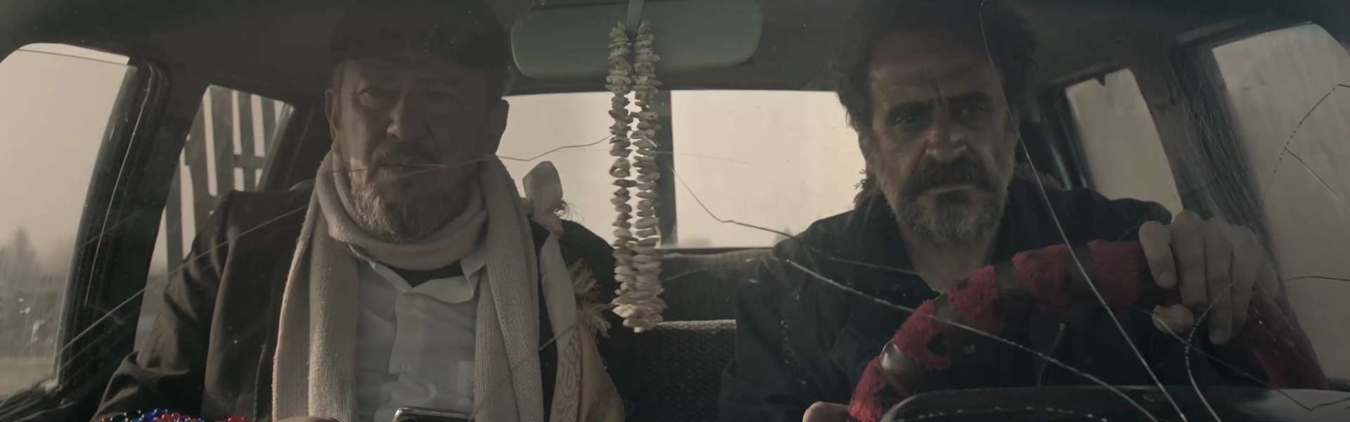 Still from Harsh Land, Distant Sky: two men sit in the front of a vehicle, one in a pale scarf and one gripping the steering wheel.