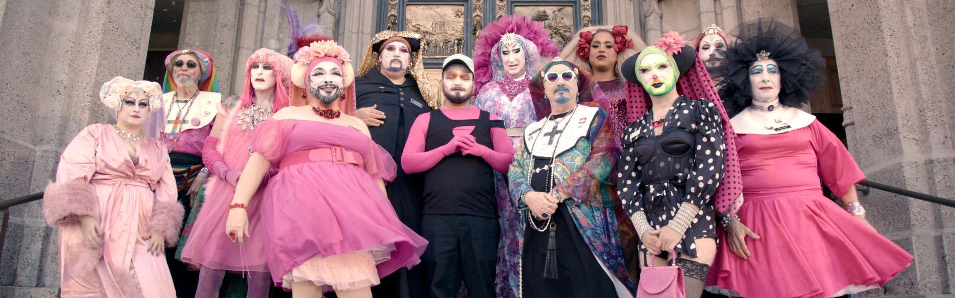 Still from Hunky Jesus: a group of people in bright pink, pale and black costumes and dramatic make-up pose together on the steps of a stone church.
