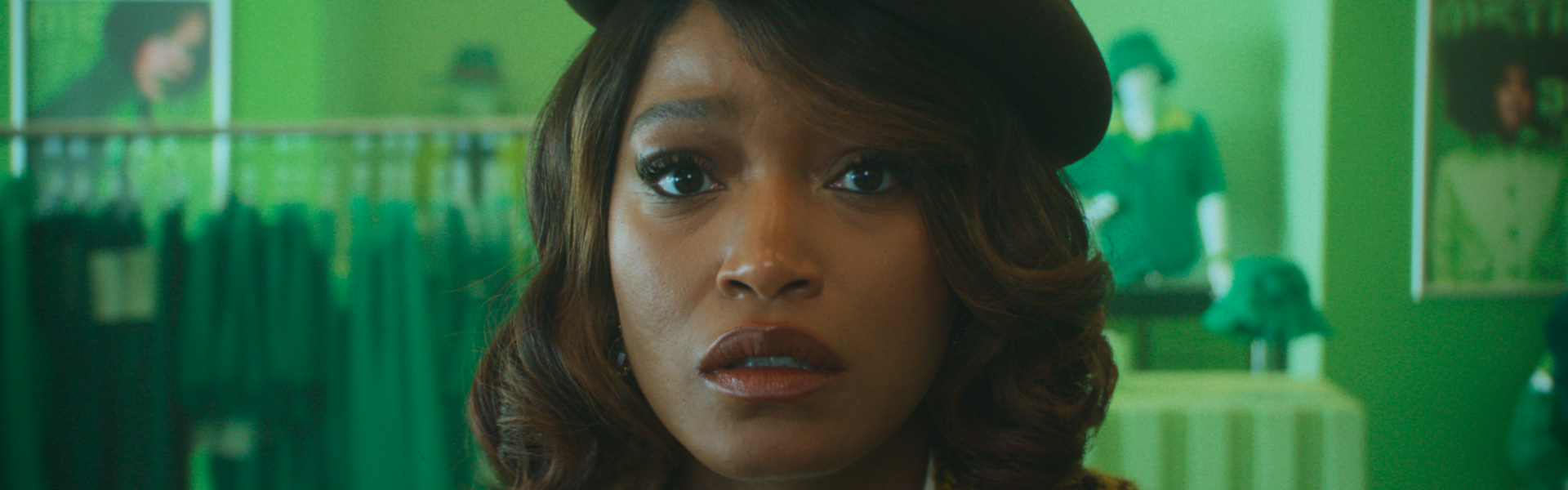 Still from I Love Boosters: a woman in a brown beret and tweed jacket looks worried, in a clothes shop lit green.