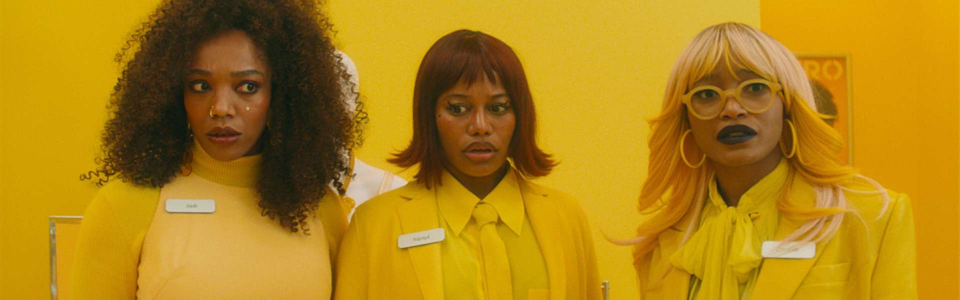 Still from I Love Boosters: three women in matching yellow jackets with name badges stand side by side against a yellow wall.
