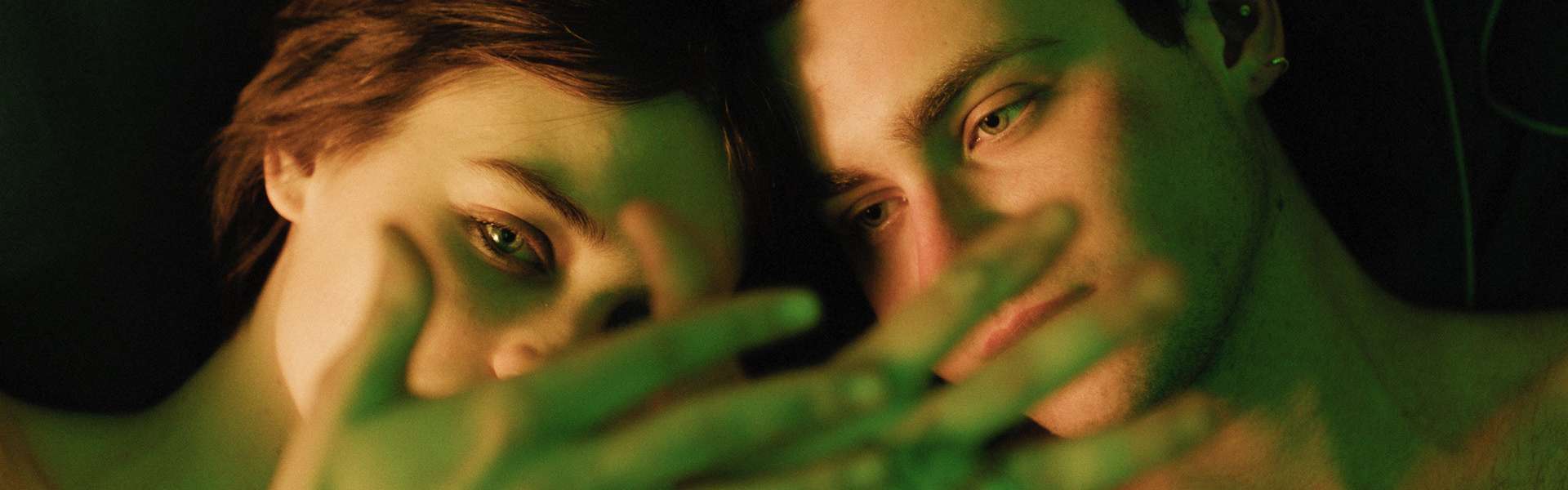 Still from I Rarely Wake Up Dreaming: two young men press their faces together with a hand held across their mouths, in green light.