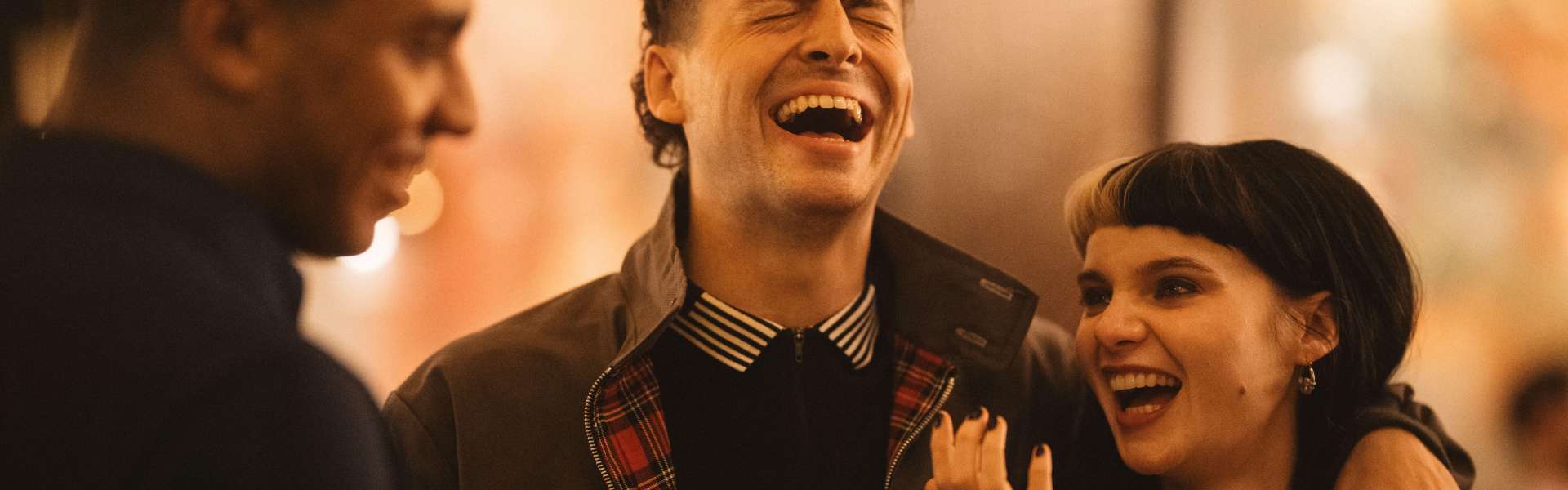 Still from I See Buildings Fall Like Lightning: a young man laughs with his eyes closed beside a young woman with a dark bob who is also laughing, with another man in profile at the left.
