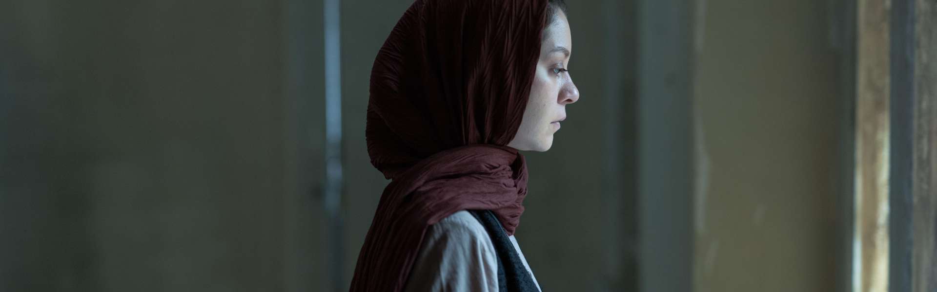 Still from It Means Hope: a woman in a red headscarf seen in profile in a dim interior.