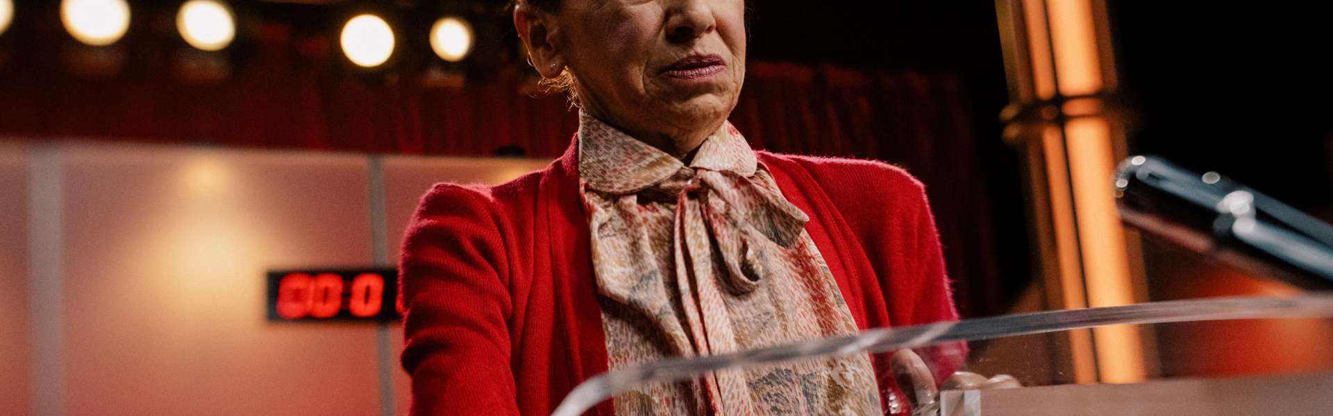 Still from Jasmine. Home. Mother.: a woman in a red jacket and ruffled cream blouse stands at a clear podium under bright stage lights, looking to one side with a tense expression.