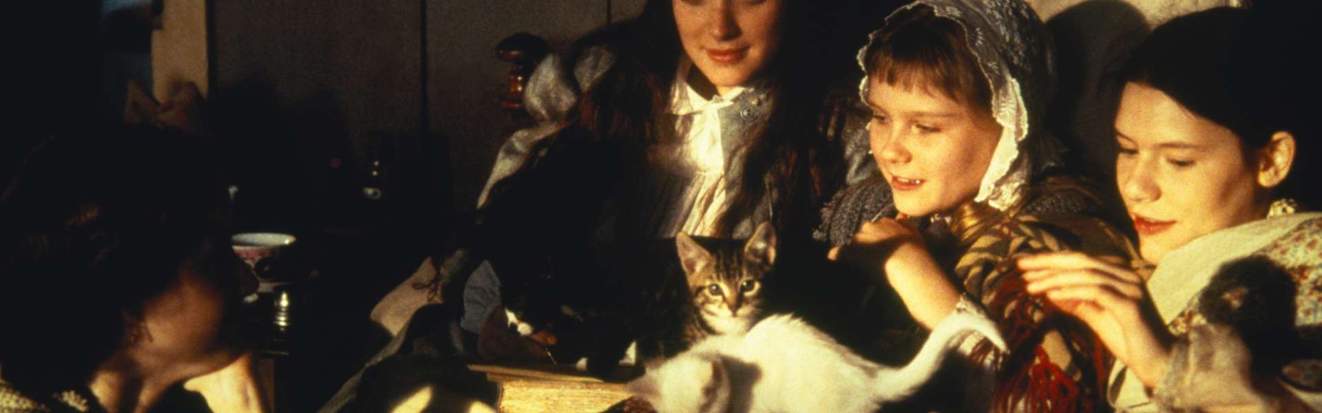 Still from Little Women: several girls in nightclothes and headscarves lie in bed, one with a white cat, under a patchwork quilt.