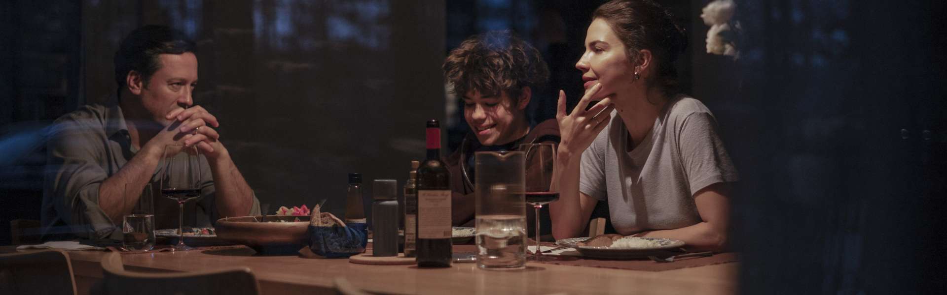 Still from Minotaur: four people sit around a dinner table in a dimly lit room, seen from outside the window.