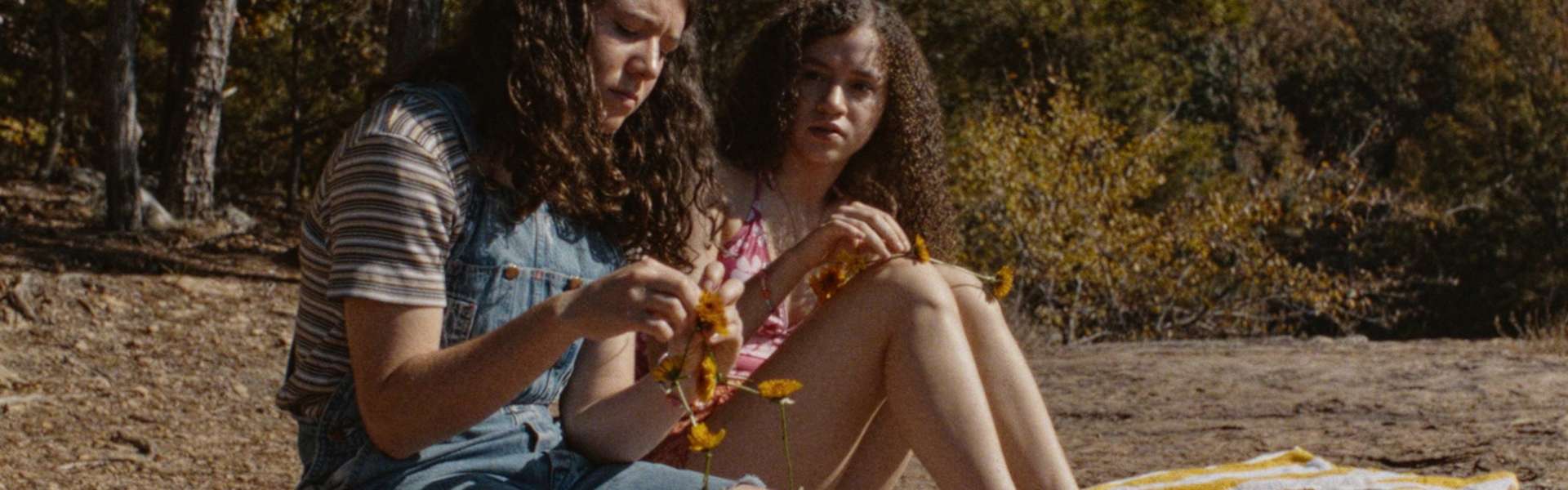 Still from Mouse: two girls with curly hair sit on rocks in a wood, one in a denim dress, both looking down at something in their hands.