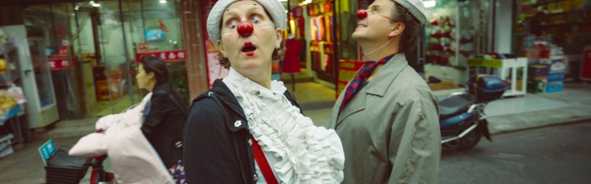 Still from My Family and Other Clowns: two people in clown costume, one with a red nose and a white ruffled shirt and the other in a hat and green coat, stand looking up on a busy street with shop signs behind them.