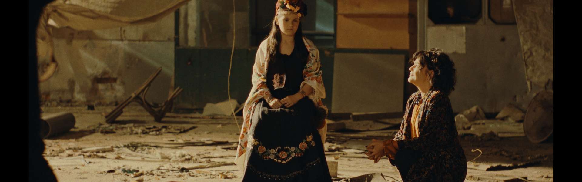 Still from Nina Roza: a woman in a long embroidered dress sits on a cloth-covered ground beside a kneeling man, in front of a wooden caravan.