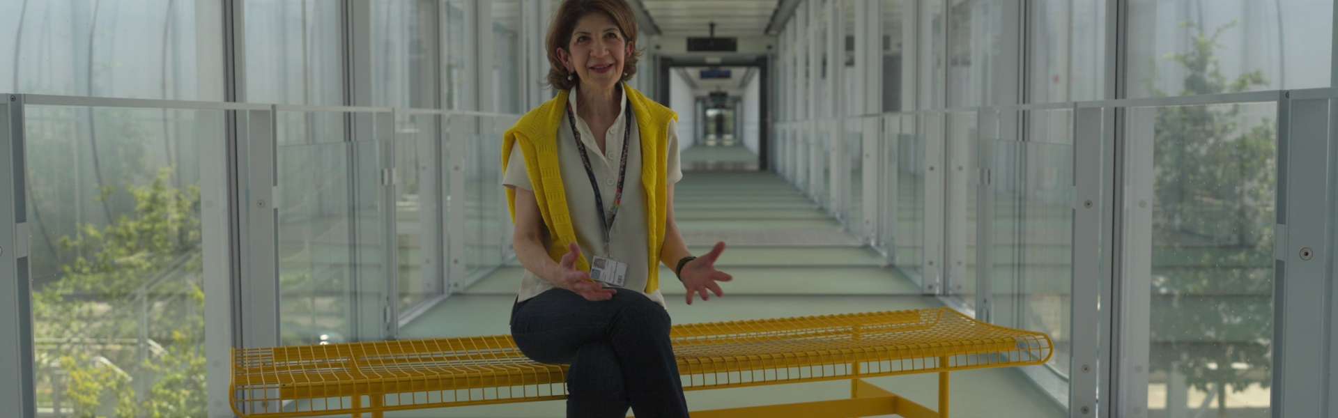Still from The Peace Particle: a woman in a yellow high-visibility vest sits on a yellow bench in a glass-walled corridor with plants outside.