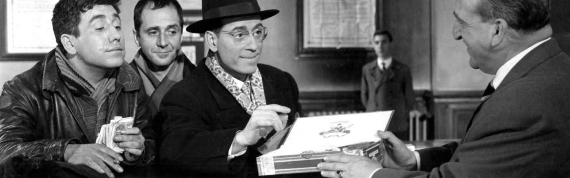 Black-and-white still from Placido: a group of men in jackets lean on an office counter, one in a hat holding a white box, as a man in a suit faces them.