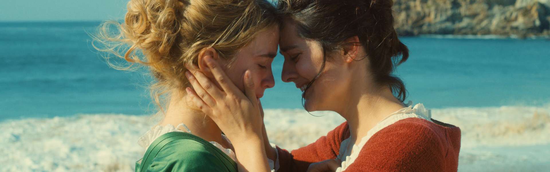 Still from Portrait of a Lady on Fire: two women in period dress, one in green and one in red, rest their foreheads together and hold each other on a beach, with cliffs behind them.
