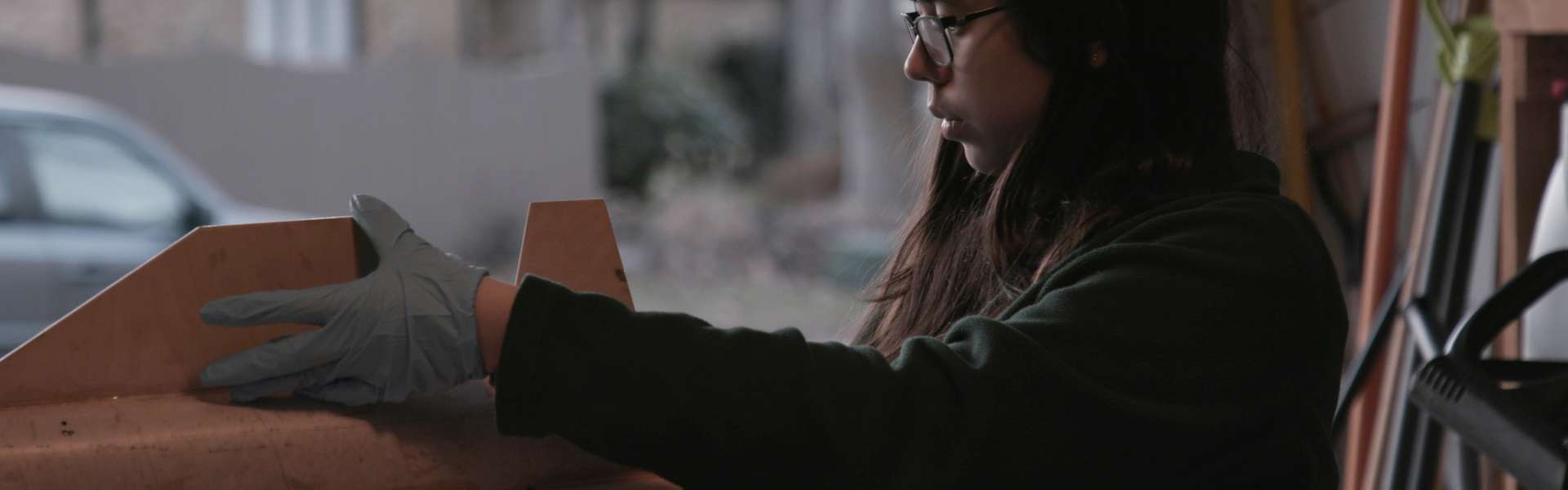 Still from Rocket Girl: a woman in glasses, seen in profile, works at a workbench with a gloved hand on a cardboard tube.