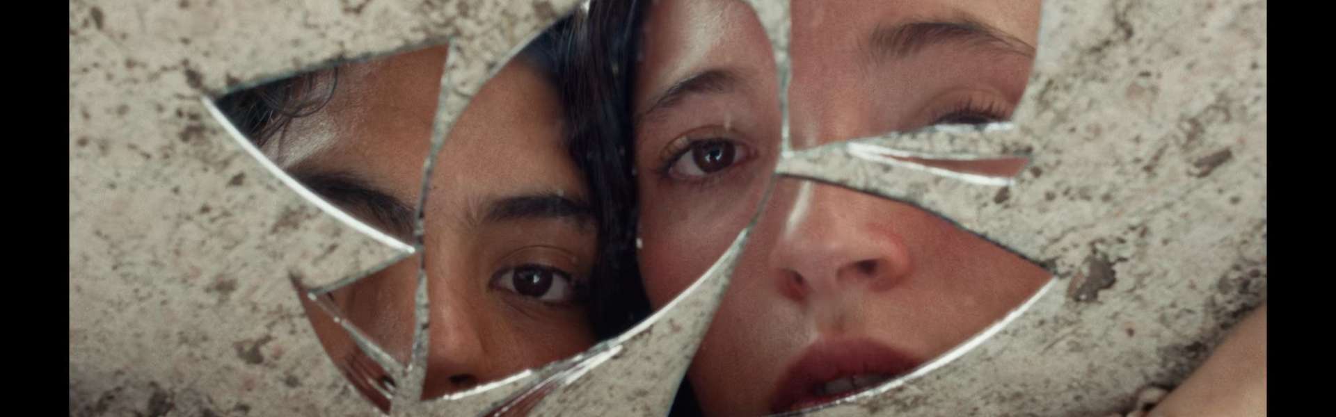 Still from Sad Girlz: a cracked mirror shows fragments of a young woman's face against a pale stone wall.