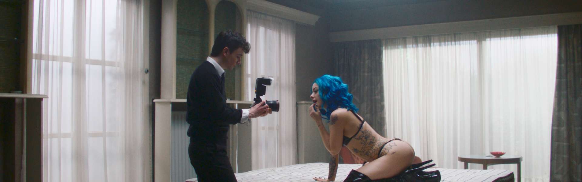 Still from Truly Naked: a camera operator films a person with blue hair kneeling on a mattress in a large room with tall windows.