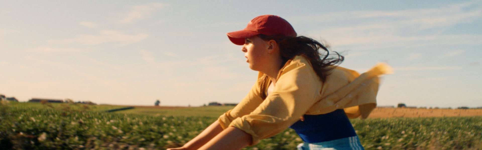 Still from The Harvest: a girl in a red cap and yellow top rides a bicycle fast across a field, her hair blowing back.