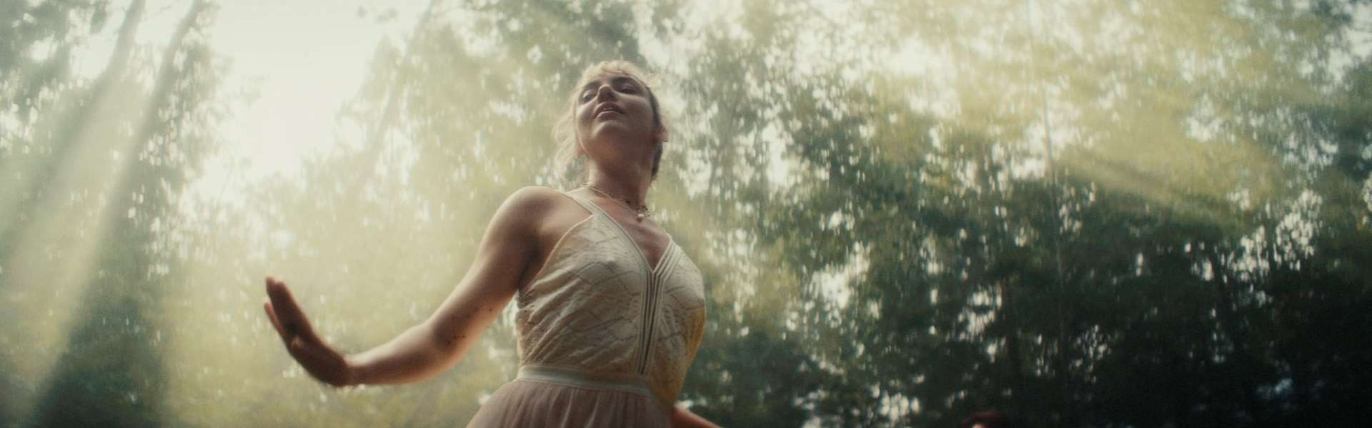Still from Todd and The Toad: a woman in a pale dress stands with her arms outstretched in a sunlit forest.
